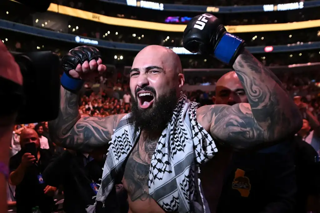 Tattooed bald UFC fighter with a beard raises his gloved fists in victory on the arena floor, a patterned towel over his shoulders and cheering crowd behind him.