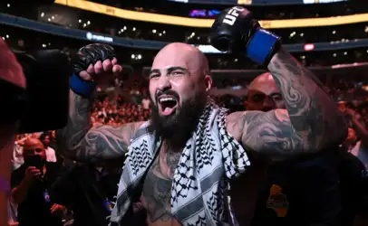 Tattooed bald UFC fighter with a beard raises his gloved fists in victory on the arena floor, a patterned towel over his shoulders and cheering crowd behind him.