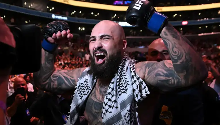 Tattooed bald UFC fighter with a beard raises his gloved fists in victory on the arena floor, a patterned towel over his shoulders and cheering crowd behind him.