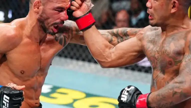 Two shirtless UFC fighters clash in the octagon; one punches the other, blood smeared on the face and a determined crowd in the background.