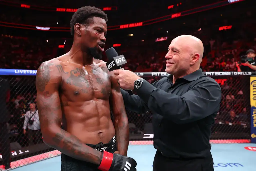 Post-fight UFC interview: a shirtless, tattooed fighter speaks into a microphone held by a man in a black shirt inside the octagon.