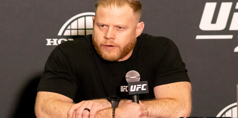 Male fighter with blond hair and beard sits at a press conference table, holding a UFC microphone.