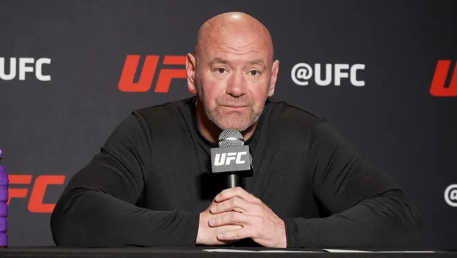 A bald man in a black shirt sits at a table holding a UFC-branded microphone during a press conference.