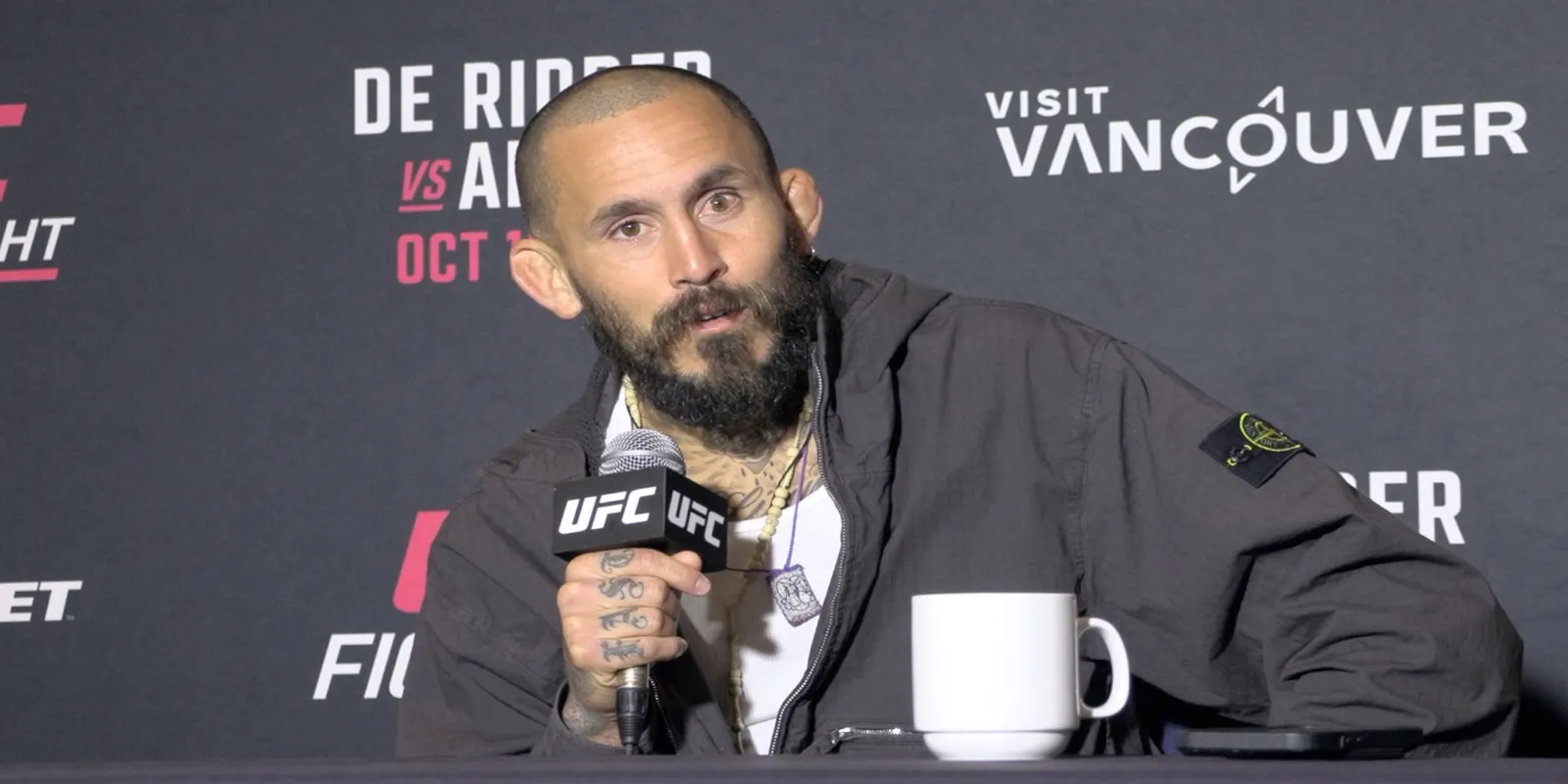 Bearded man in a dark jacket speaks into a UFC-branded microphone at a press event, with a white mug on the table in front of him.