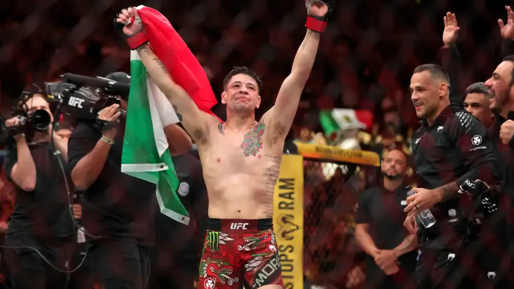 UFC fighter raises arms in victory, holding a Mexican flag inside the cage while teammates cheer nearby.