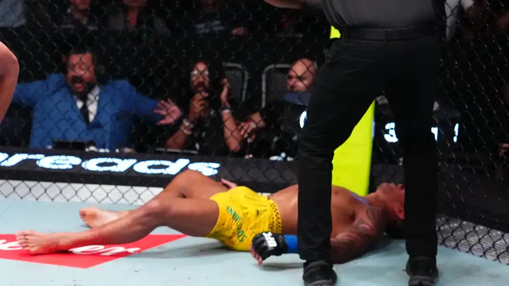 Fighter in yellow shorts lies on the mat inside a fenced MMA cage while a referee stands nearby and spectators watch in the background.