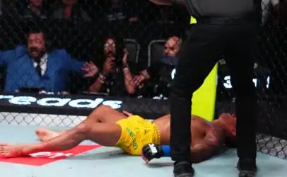 Fighter in yellow shorts lies on the mat inside a fenced MMA cage while a referee stands nearby and spectators watch in the background.