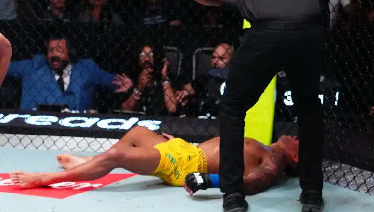 Fighter in yellow shorts lies on the mat inside a fenced MMA cage while a referee stands nearby and spectators watch in the background.