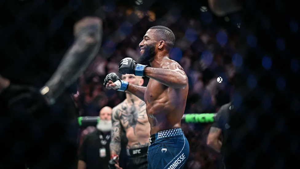 UFC fighter in blue shorts celebrating with fists up inside the octagon, crowd in background.