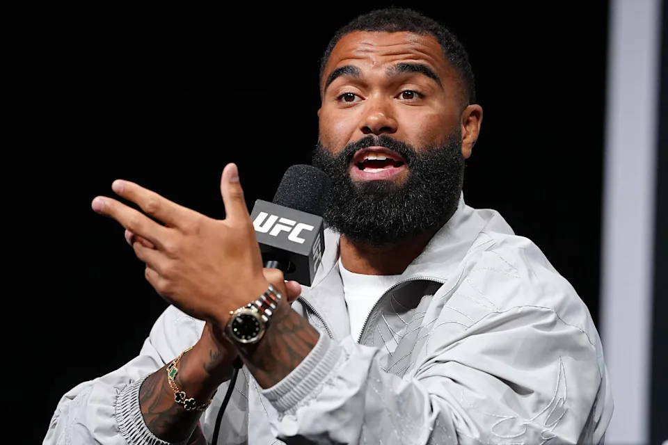 Bearded man speaks into a UFC-branded microphone, gesturing with his hands during a press event.