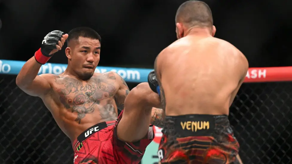 9a6b3c90-b4b7-11f1-bca0-c1ba24c9211cjpg - AfroCombat Two UFC fighters clash in the octagon as one delivers a kick to the other's midsection, both wearing gloves and shorts.