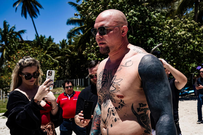 Shirtless tattooed man wearing sunglasses at a sunny beach as onlookers photograph him, palm trees and blue sky in the background.