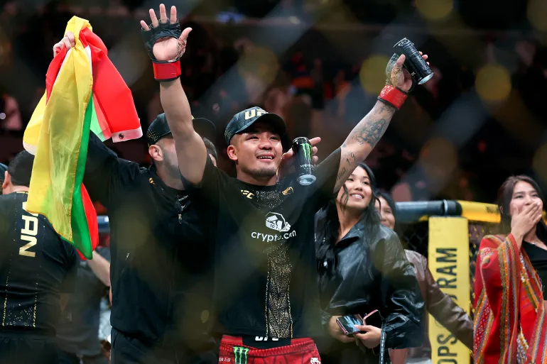 AFP__20260920__2296162547__v1__HighRes__Ufc331VanVPantoja-1789882334 - AfroCombat MMA fighter in black shirt and red shorts raises arms in victory, holding a drink can and a tri-color flag as fans cheer in the arena.
