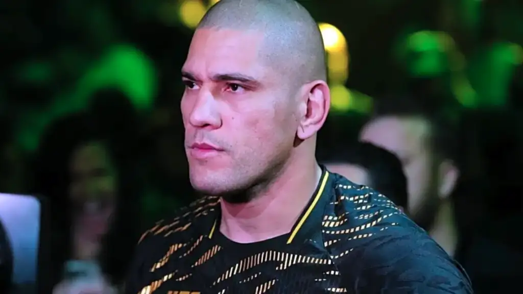 Man with a shaved head and light stubble, wearing a black and gold patterned shirt, at a crowded event with green stage lights in the background.