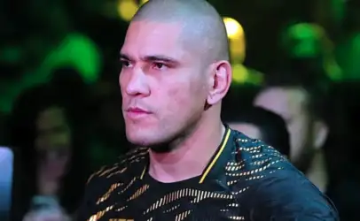 Man with a shaved head and light stubble, wearing a black and gold patterned shirt, at a crowded event with green stage lights in the background.