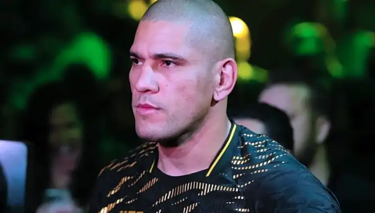 Man with a shaved head and light stubble, wearing a black and gold patterned shirt, at a crowded event with green stage lights in the background.