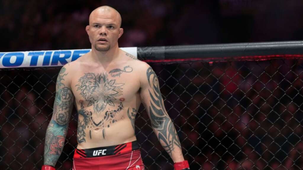 Bald, tattooed MMA fighter standing inside a UFC cage, wearing red shorts and gloves, looking forward.