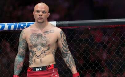 Bald, tattooed MMA fighter standing inside a UFC cage, wearing red shorts and gloves, looking forward.