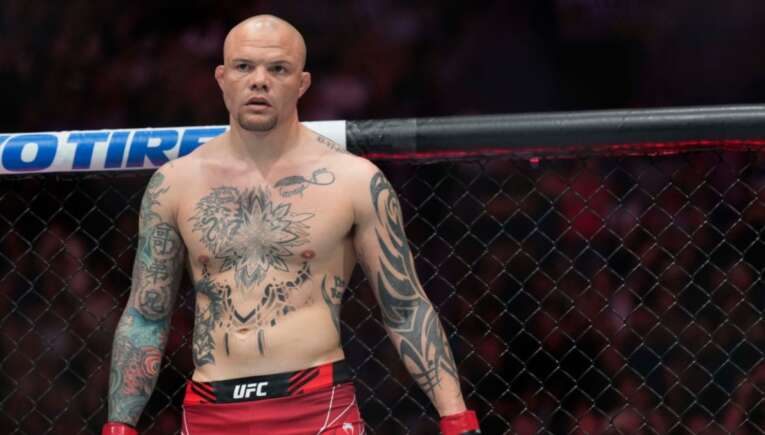 Bald, tattooed MMA fighter standing inside a UFC cage, wearing red shorts and gloves, looking forward.