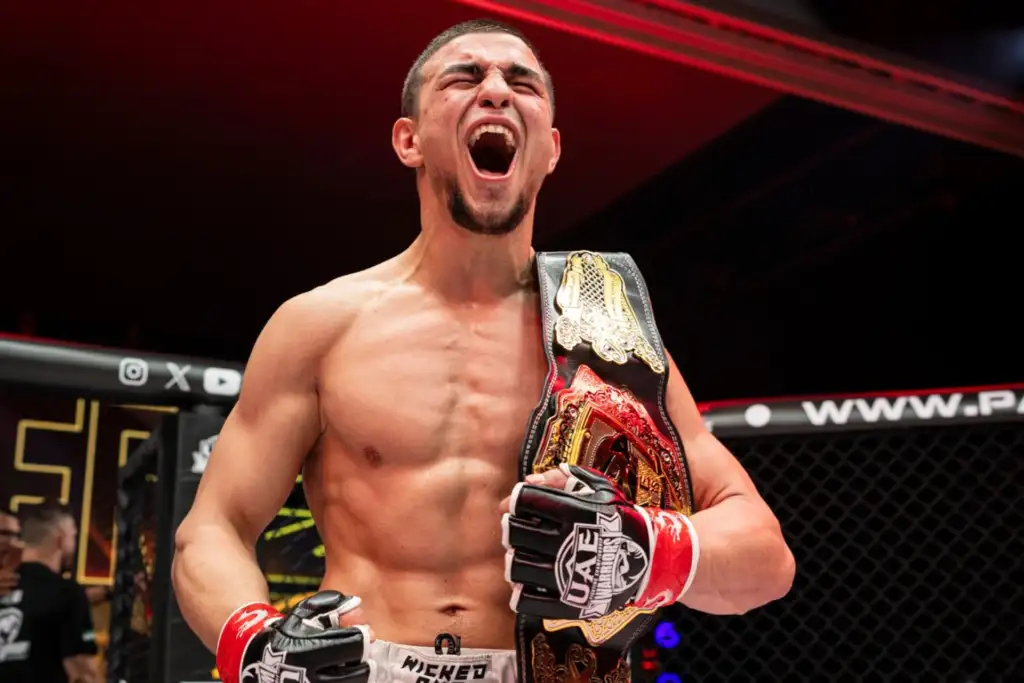 MMA fighter shirtless in a cage, shouting triumphantly while holding a gold championship belt over his shoulder