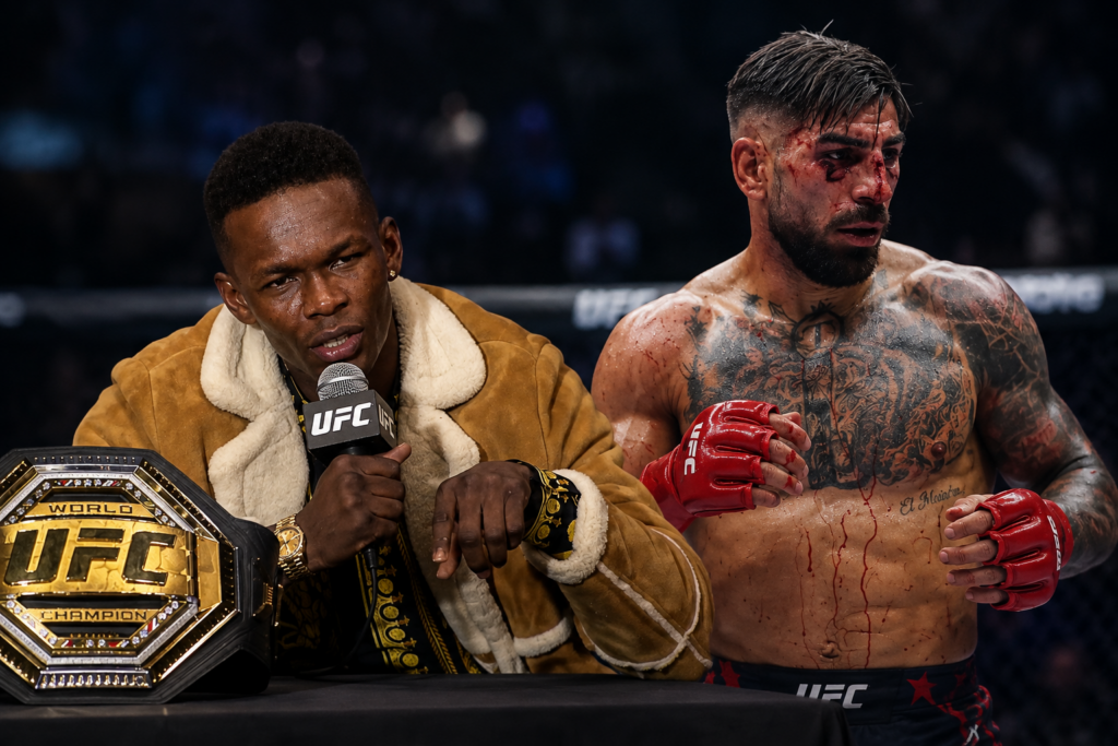 UFC post-fight interview: a man in a brown shearling coat speaks into a mic beside the UFC belt on the table, while a bloodied, tattooed fighter stands with red gloves nearby.