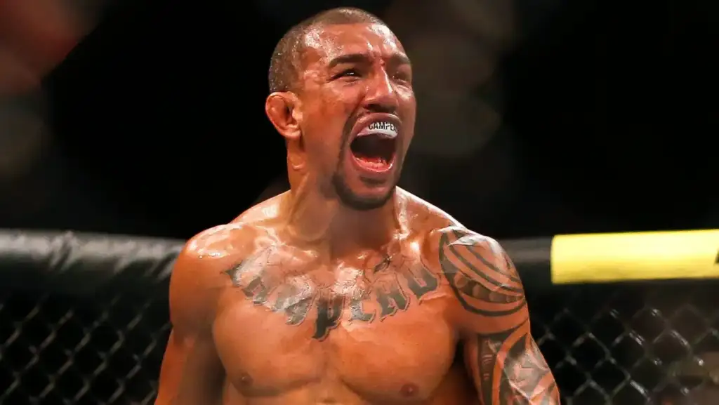 MMA fighter in an octagon shouting, with a mouthguard visible and sweat on his upper body, tattoos on his chest and arm.