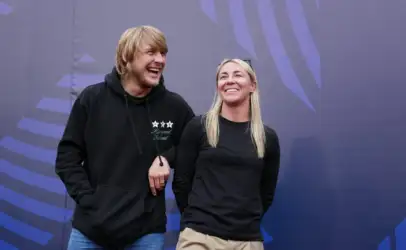 Two smiling people stand side by side with linked arms against a purple geometric backdrop.