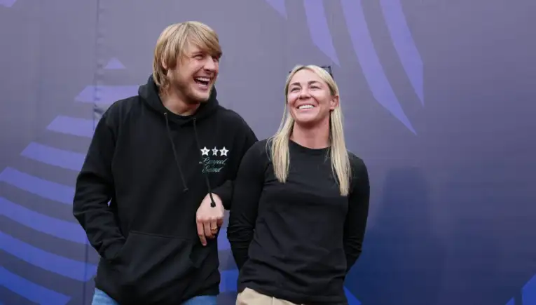Two smiling people stand side by side with linked arms against a purple geometric backdrop.