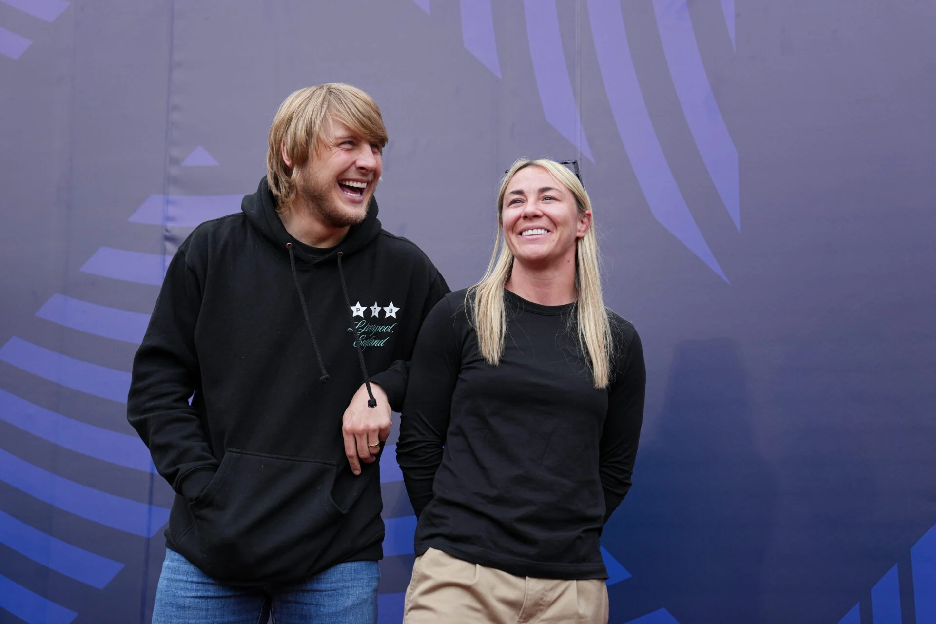 Two smiling people stand side by side with linked arms against a purple geometric backdrop.