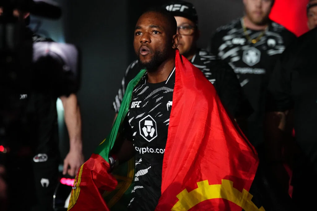 UFC fighter walks toward the arena with a red-and-green flag draped over his shoulders, wearing black gear with white logos and a focused expression.