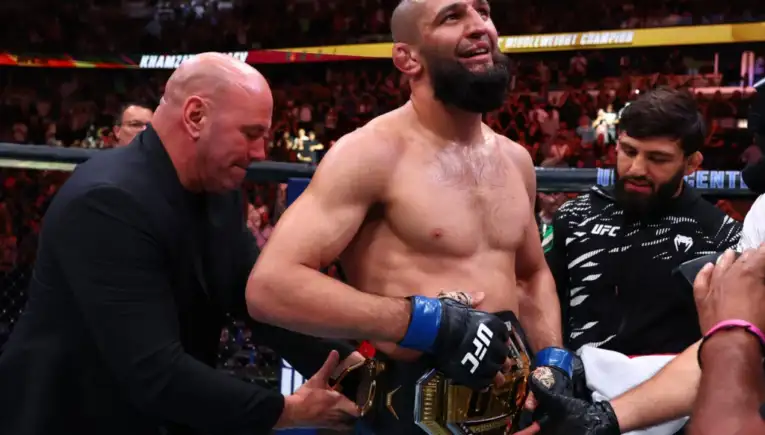 UFC fighter with a championship belt around his waist stands in the octagon after victory, surrounded by teammates adjusting the belt.