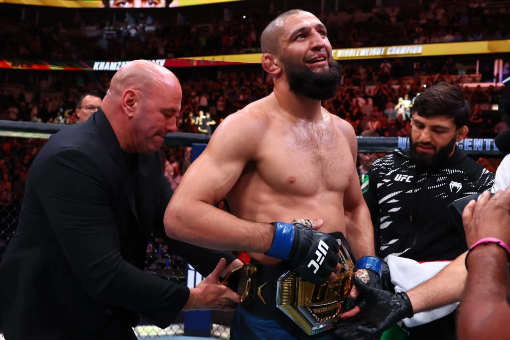 UFC fighter with a championship belt around his waist stands in the octagon after victory, surrounded by teammates adjusting the belt.
