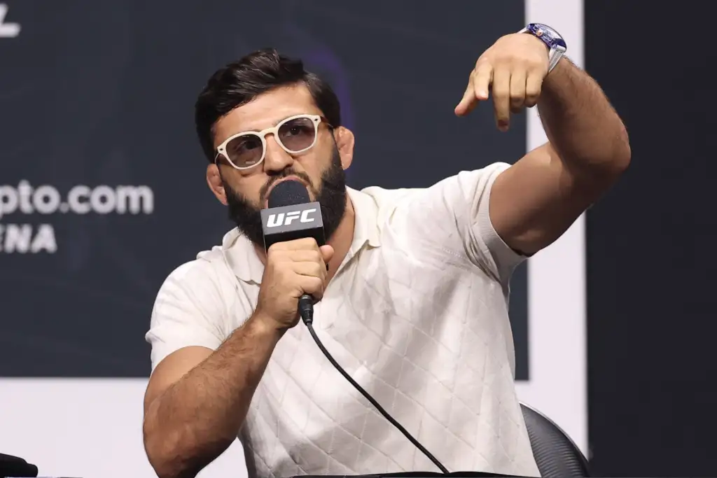 Man at a press event speaks into a UFC microphone while gesturing with his right arm raised and pointing outward, wearing white sunglasses and a light polo shirt.