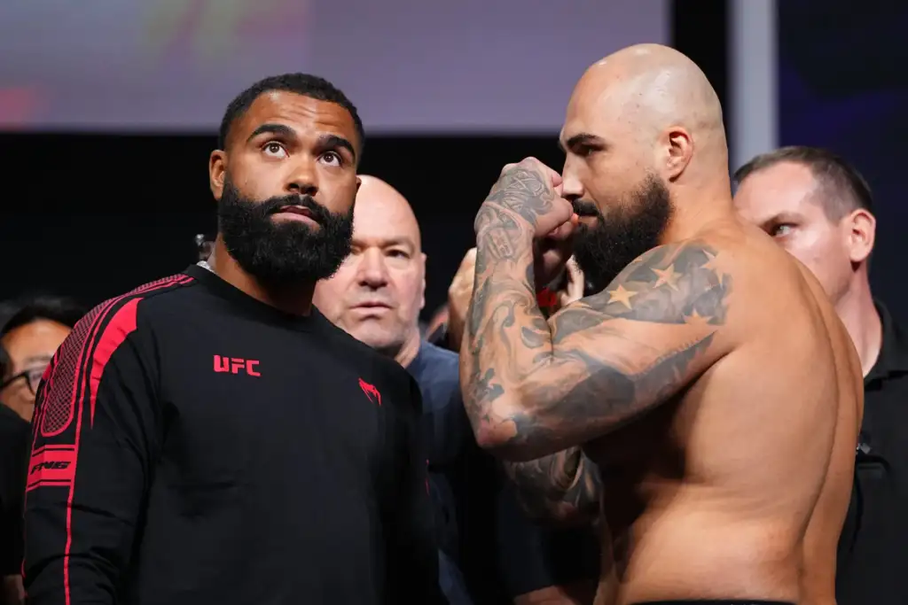 Two muscular men facing off at a UFC event: one in a black UFC shirt, the other shirtless with tattoos, as crowd watches in the background.
