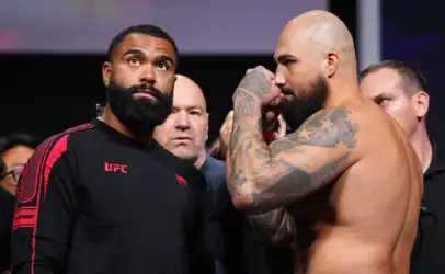 Two muscular men facing off at a UFC event: one in a black UFC shirt, the other shirtless with tattoos, as crowd watches in the background.