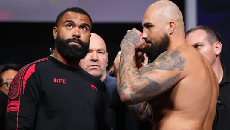 Two muscular men facing off at a UFC event: one in a black UFC shirt, the other shirtless with tattoos, as crowd watches in the background.