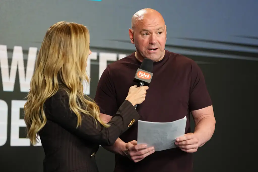 Man with a shaved head being interviewed on stage, speaking into a handheld microphone held by a woman with long blonde hair and notes in hand.