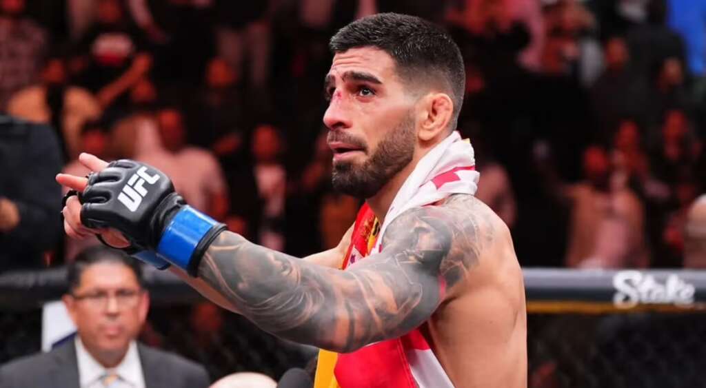 UFC fighter with a blue glove, tattoos, and a flag draped around his shoulders in a packed arena, raising a fist in celebration.
