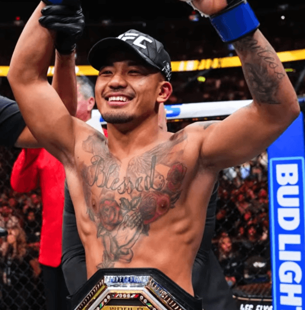 UFC fighter with arms raised in victory inside the octagon, wearing a cap and a championship belt, tattoos on the chest.