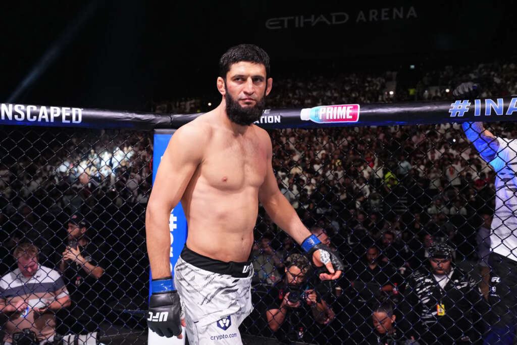 Bearded male MMA fighter shirtless inside the UFC octagon, wearing blue gloves and white shorts, looking at the camera with a crowded arena behind him.