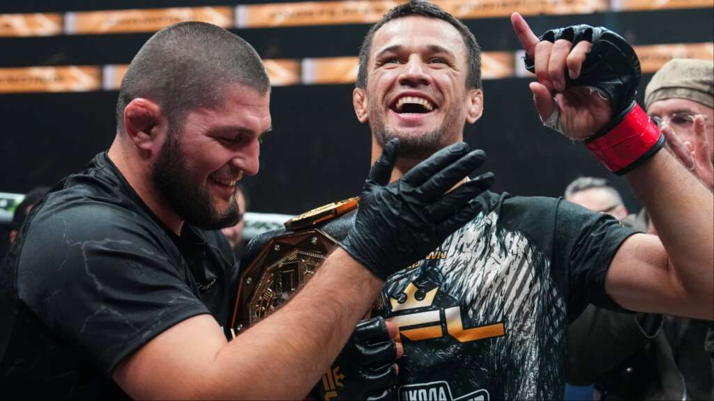 Two victorious fighters celebrate together, one wearing a championship belt and smiling broadly.