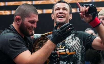 Two victorious fighters celebrate together, one wearing a championship belt and smiling broadly.