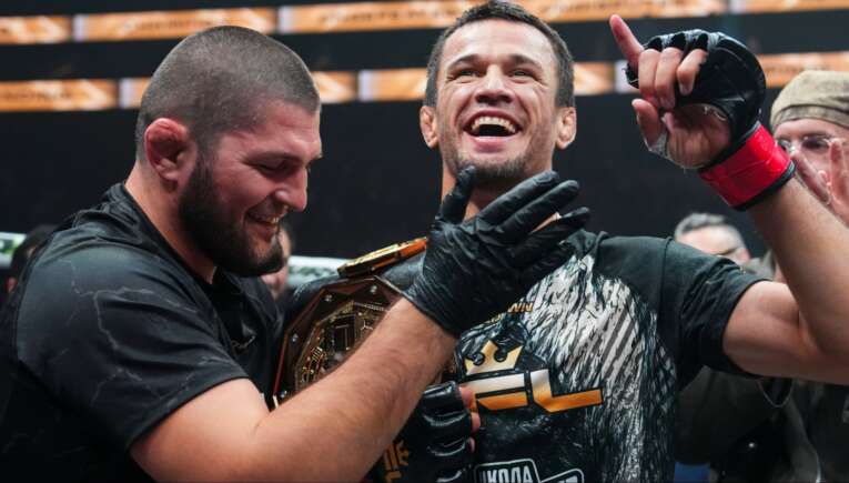 Two victorious fighters celebrate together, one wearing a championship belt and smiling broadly.