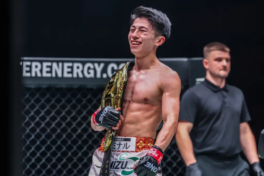 Smiling male mixed martial artist, shirtless, with a gold championship belt draped over his shoulder inside an MMA cage.