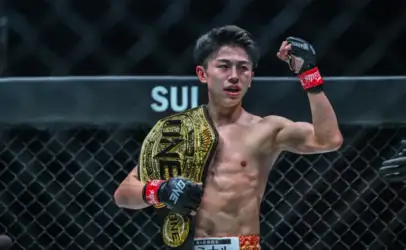 Male mixed martial artist posing in a cage with a gold championship belt over his shoulder; ONE Championship logo visible on belt and gloves