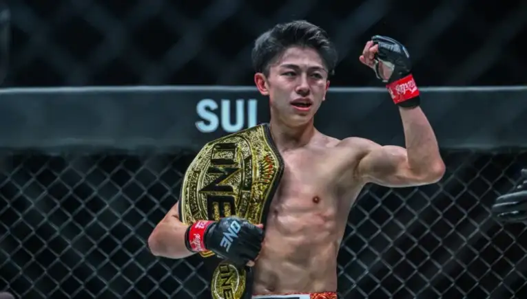 Male mixed martial artist posing in a cage with a gold championship belt over his shoulder; ONE Championship logo visible on belt and gloves