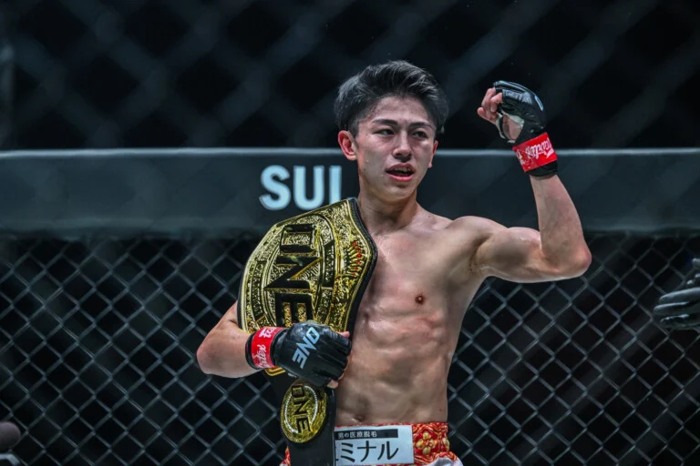 Male mixed martial artist posing in a cage with a gold championship belt over his shoulder; ONE Championship logo visible on belt and gloves