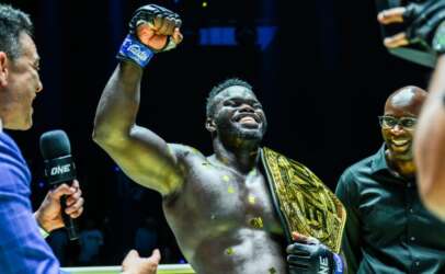 Black muscular fighter raises his fist in victory, wearing a gold championship belt during a post-fight interview with a interviewer holding a mic beside him.