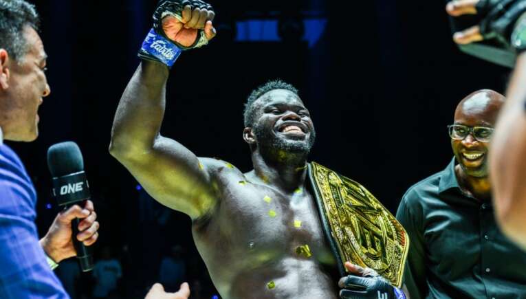 Black muscular fighter raises his fist in victory, wearing a gold championship belt during a post-fight interview with a interviewer holding a mic beside him.
