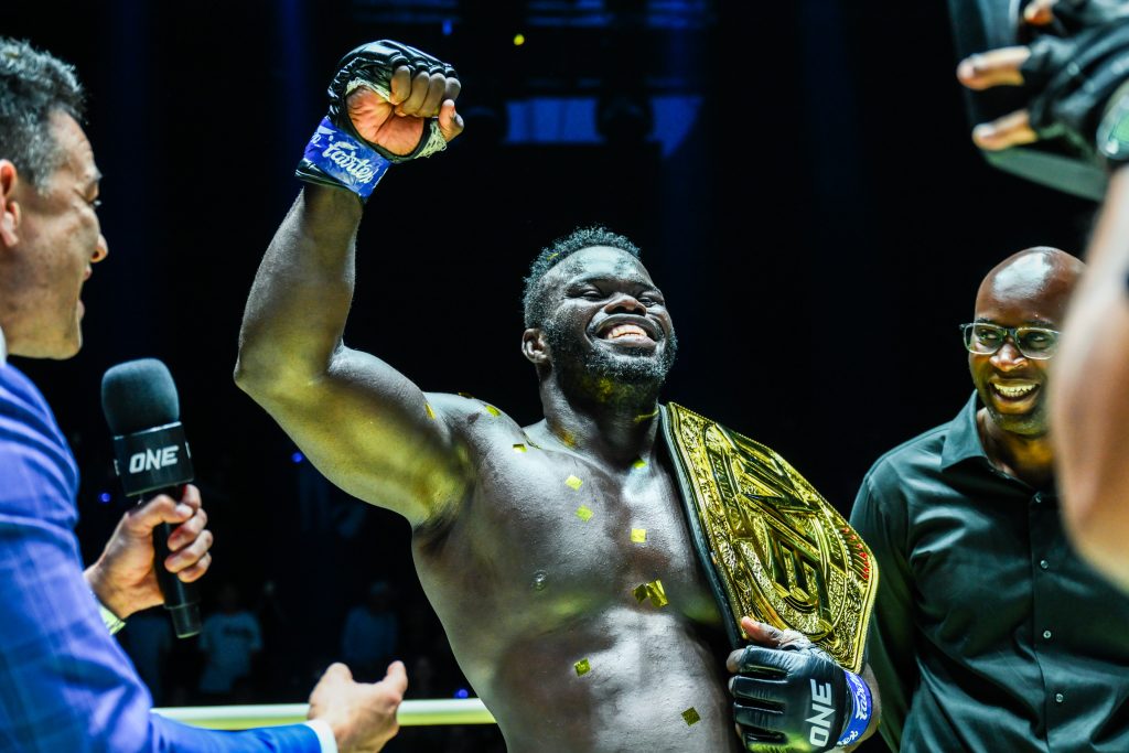 Black muscular fighter raises his fist in victory, wearing a gold championship belt during a post-fight interview with a interviewer holding a mic beside him.